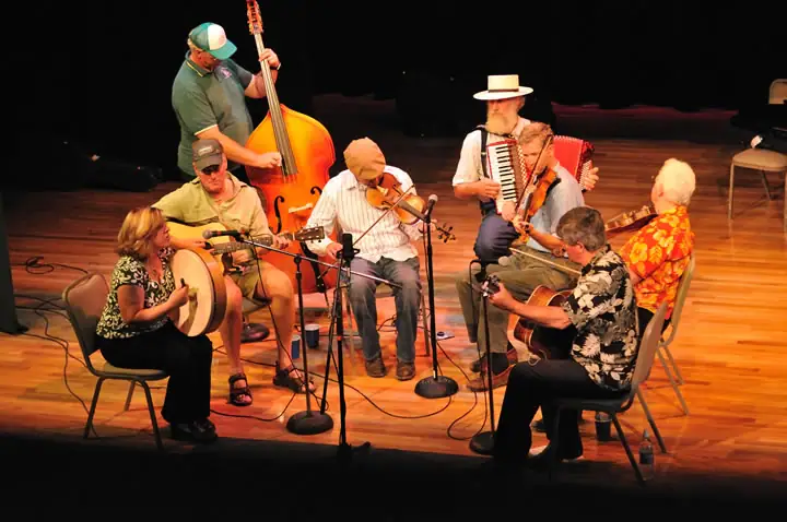 Musicians perform together on a wooden stage with string instruments, an accordion, microphones, and chairs.