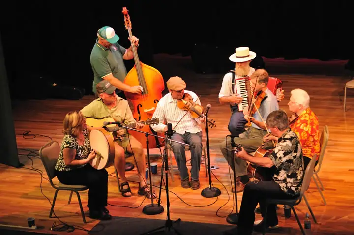 Musicians perform together on a stage with stringed instruments, accordions, a drum, microphones, and cables.