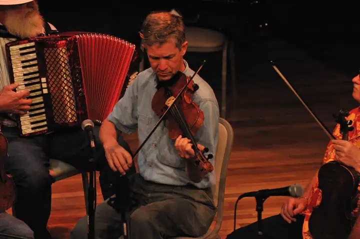 Three seated musicians perform with an accordion, violin, and bowed string instrument on a stage.