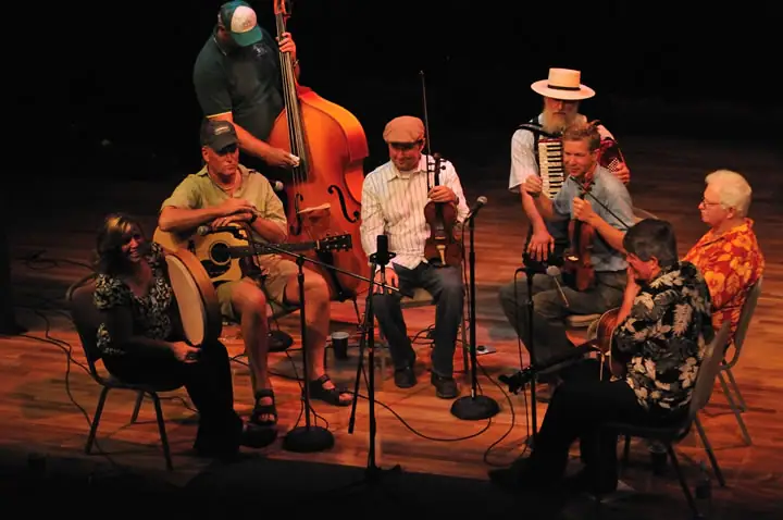 Eight musicians sit and stand on a wooden stage with string instruments and microphones.