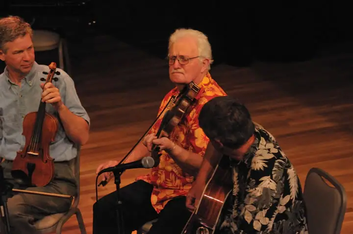 Three seated musicians play string instruments on a dimly lit wooden stage.
