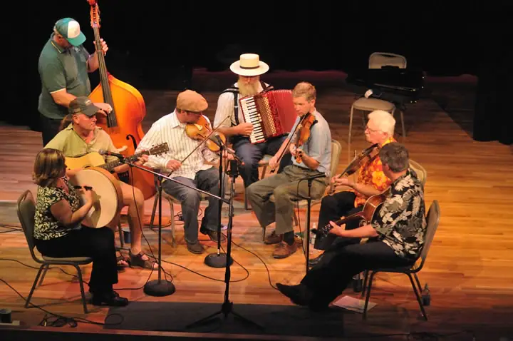 Eight musicians perform together on a wooden stage with fiddles, accordion, guitars, bass, and a hand drum.