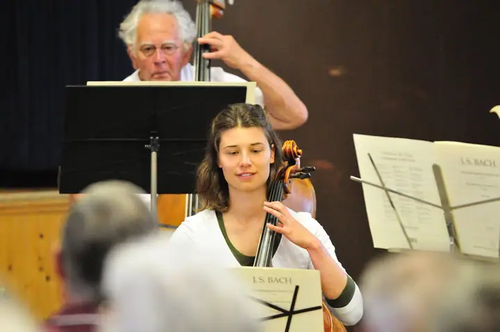 Two musicians perform string instruments behind music stands as audience members watch from the foreground.