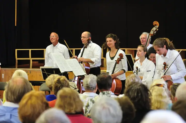 Musicians with string instruments perform before a seated audience in an indoor venue.