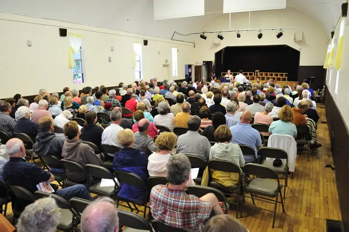 A large audience fills a hall, facing a stage where several people are seated.