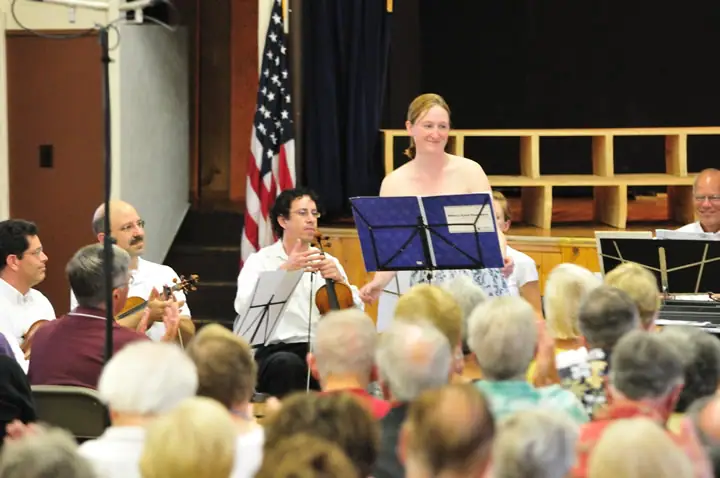 Musicians perform before a seated audience, with a standing woman at a music stand and an American flag behind them.