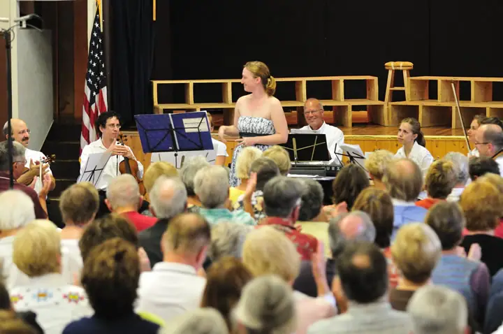 A woman stands before seated musicians while a large audience watches in an indoor performance space.
