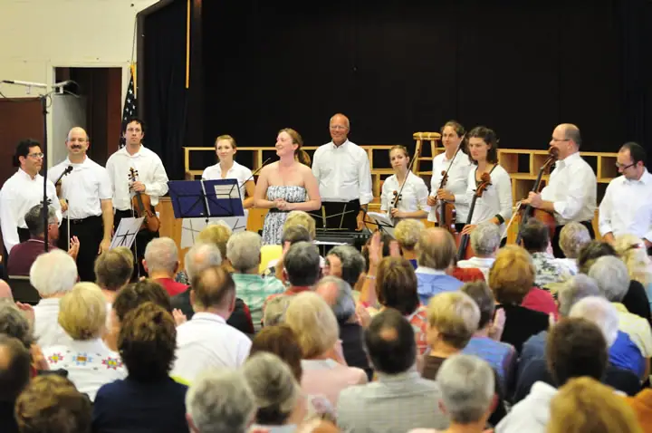 Musicians stand on an indoor stage with string instruments before a seated audience.