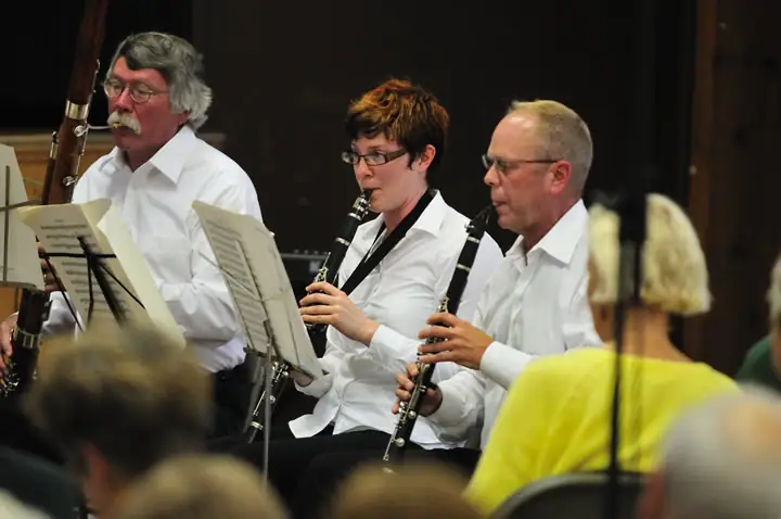 Three woodwind musicians perform from sheet music while audience members watch.
