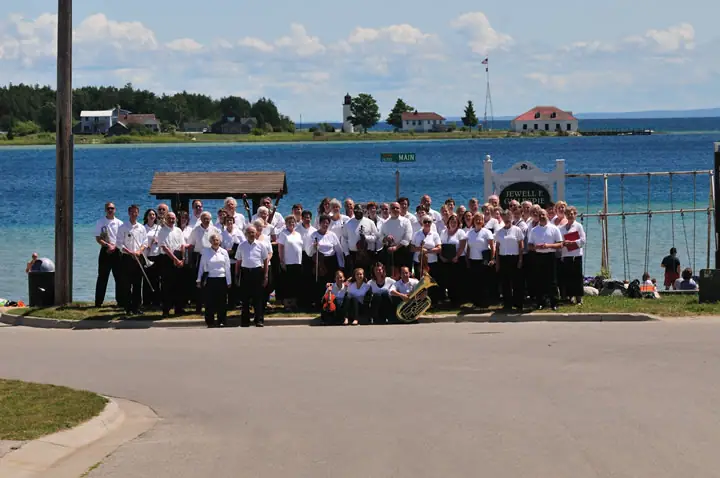 A large group of musicians poses beside the waterfront with instruments and buildings behind them.