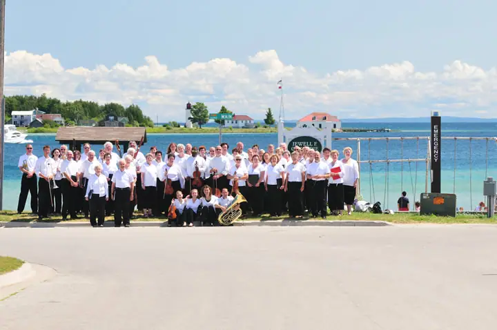 A large group of musicians poses beside the waterfront with brass instruments and buildings behind them.