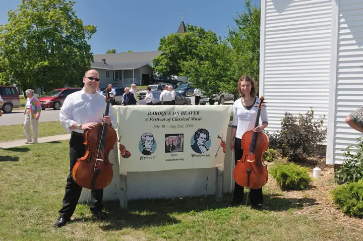 Two cellists stand beside a festival banner outside a white-sided building.