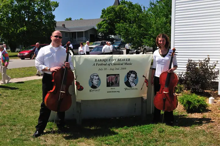 Two cellists hold a festival banner outdoors beside a house and parked cars.