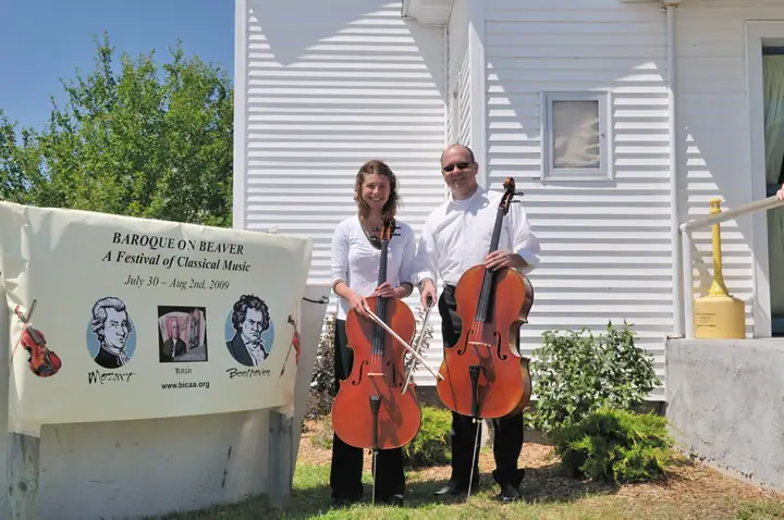 Two cellists stand beside a festival banner outside a white-sided building.