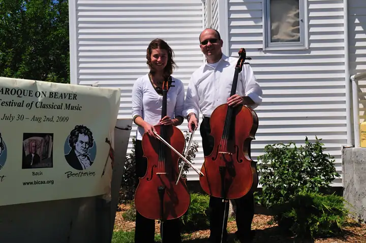 Two musicians pose outdoors with cellos beside a sign advertising a classical music festival.