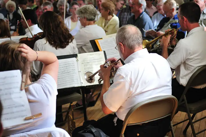 Seated musicians play brass and string instruments from sheet music in a crowded indoor ensemble.