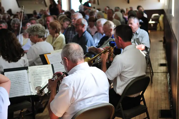 Seated brass musicians play from sheet music among an audience in a wood-paneled room.