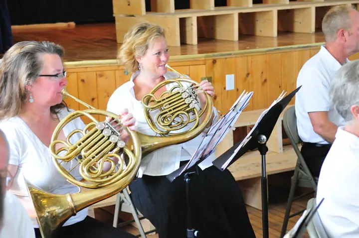 Musicians play French horns beside music stands in a wood-paneled performance room.