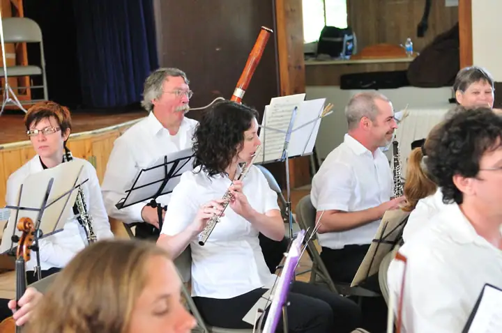 Seated musicians in white shirts play woodwind instruments beside music stands indoors.