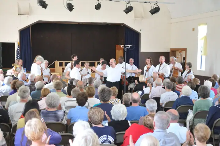 Musicians perform on a stage before a seated audience in a bright hall.