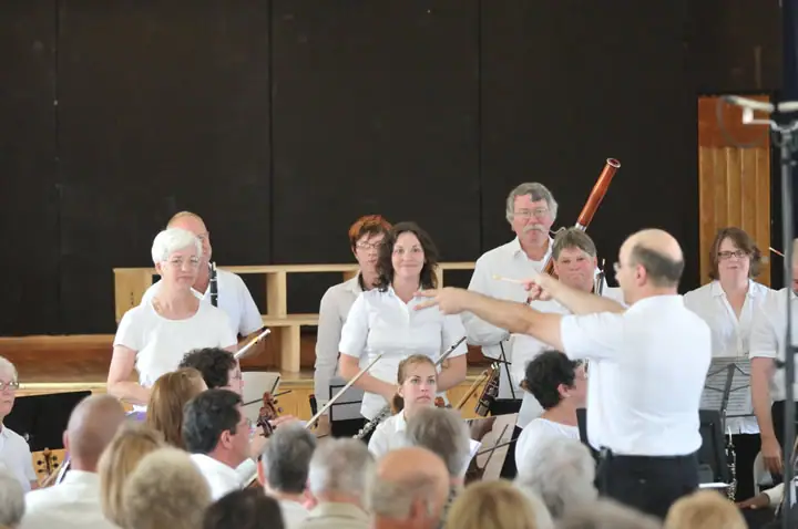A conductor leads musicians while an audience watches from the foreground.