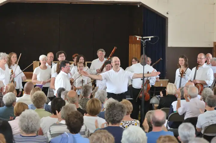 A conductor leads a string ensemble onstage before a seated audience.