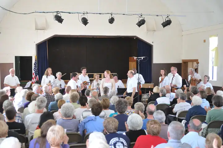Musicians perform on a stage before a seated audience in a bright hall.