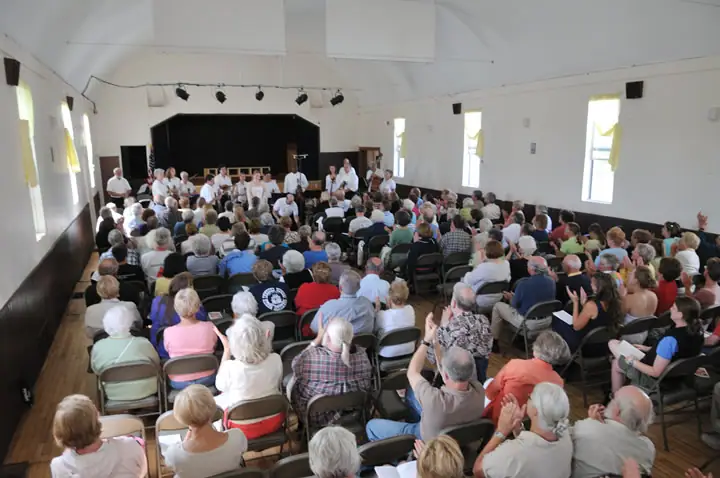 A seated audience applauds performers on a stage inside a bright hall.