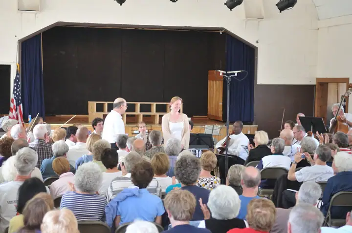 A crowded hall faces a stage where two people stand among seated musicians and instruments.