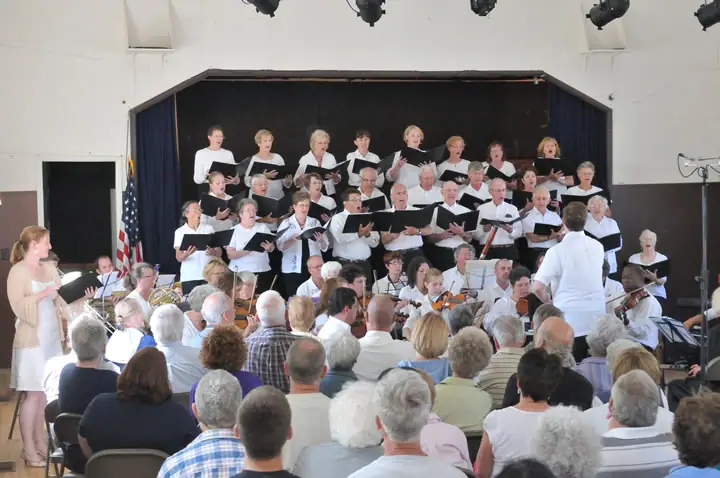 A choir performs onstage with a conductor and instrumentalists before a seated audience.