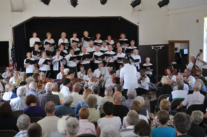 A choir and orchestra perform before a seated audience in an indoor hall.