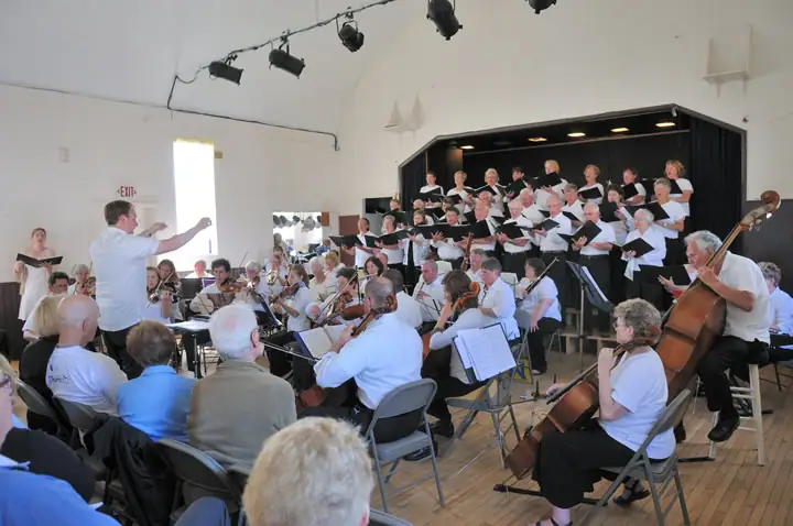 A conductor leads a choir and seated orchestra performing before an audience in a hall.