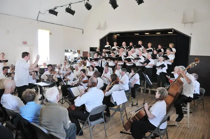 Choir and string orchestra perform indoors under the direction of a conductor.