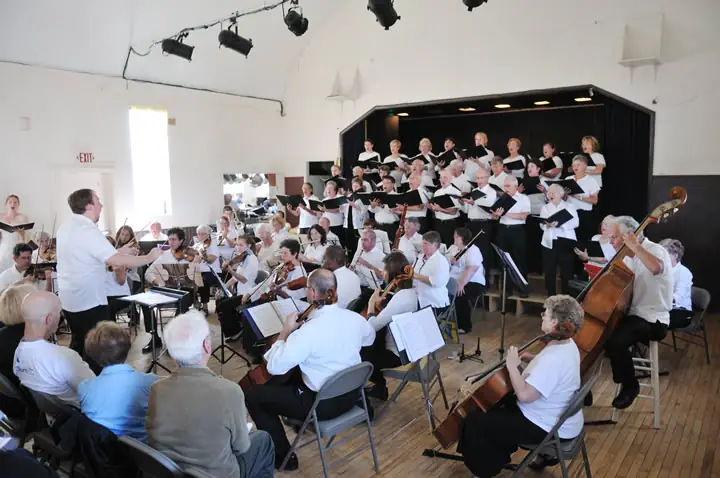A choir and orchestra perform together in a bright hall, with a conductor leading from the foreground.