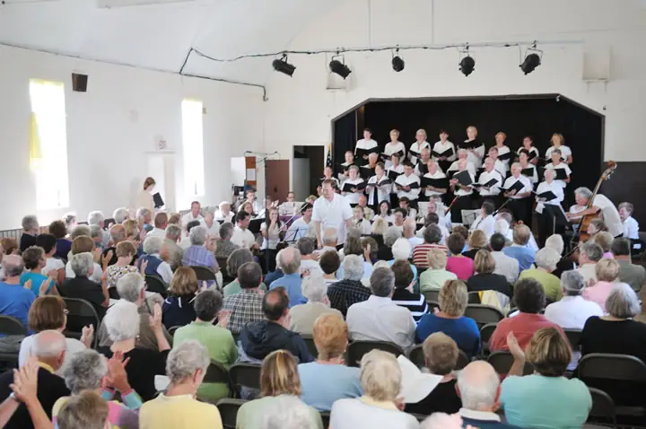 A choir and instrumental ensemble perform on a small stage before a large seated audience.