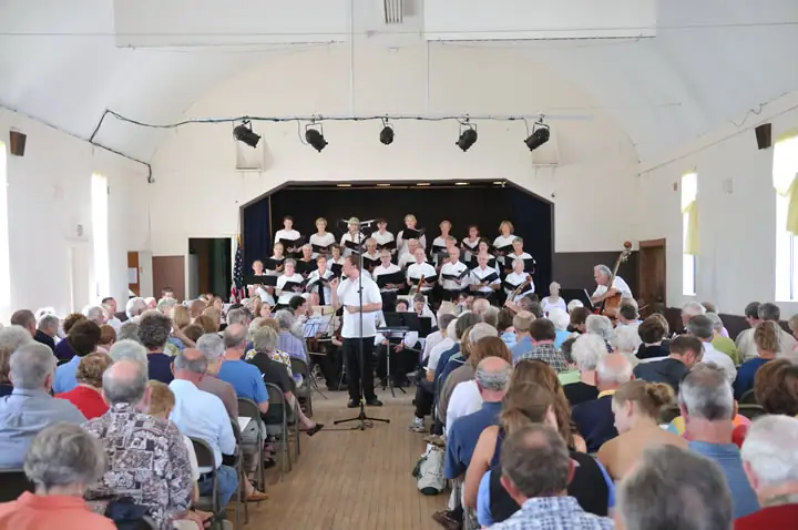 A choir performs onstage before a seated audience, with a conductor and double-bass player.