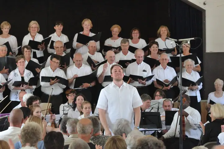 A conductor leads a choir and accompanying musicians before an audience indoors.