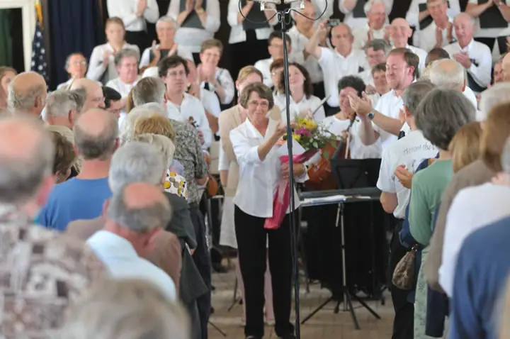 A woman holds a bouquet before a clapping choir and audience during a musical performance.
