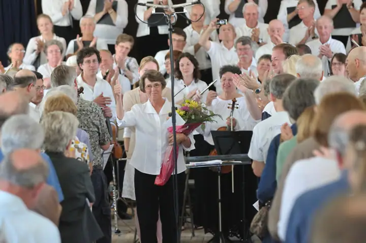 A woman holding flowers gives a thumbs-up as a crowd applauds around musicians and music equipment.