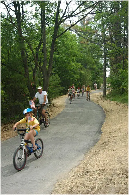 Cyclists, including a child, ride along a paved path through a wooded area while carrying small American flags.