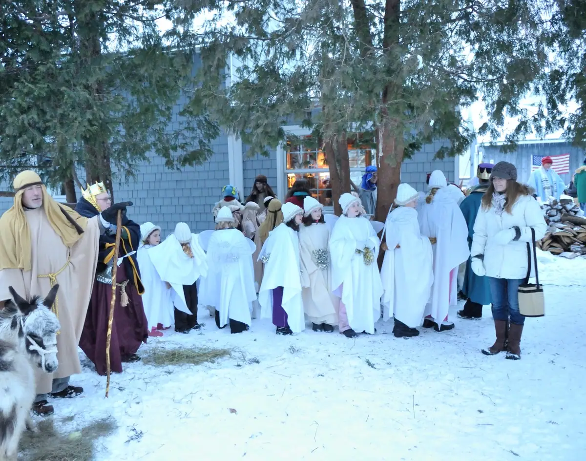 People in winter costumes gather with a donkey in a snowy yard.
