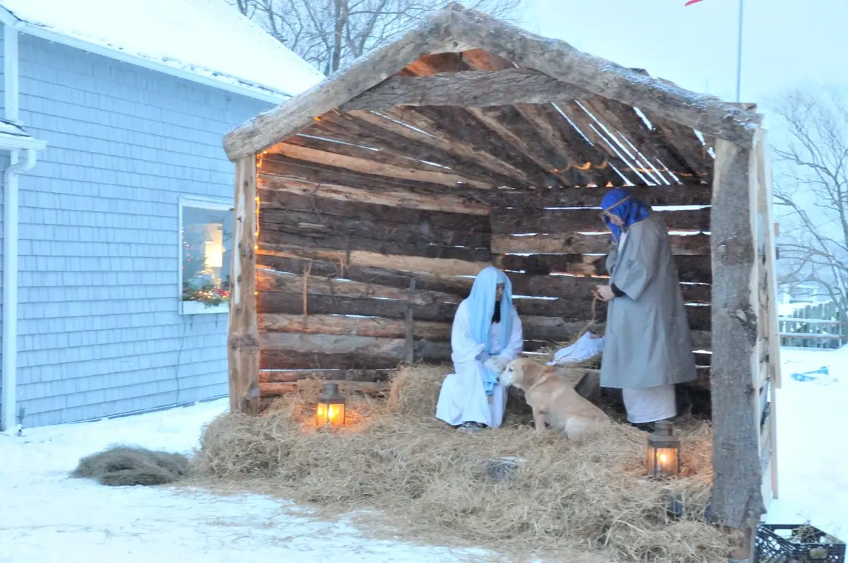 Two costumed people and a dog stand in a hay-filled wooden shelter surrounded by snow and lanterns.