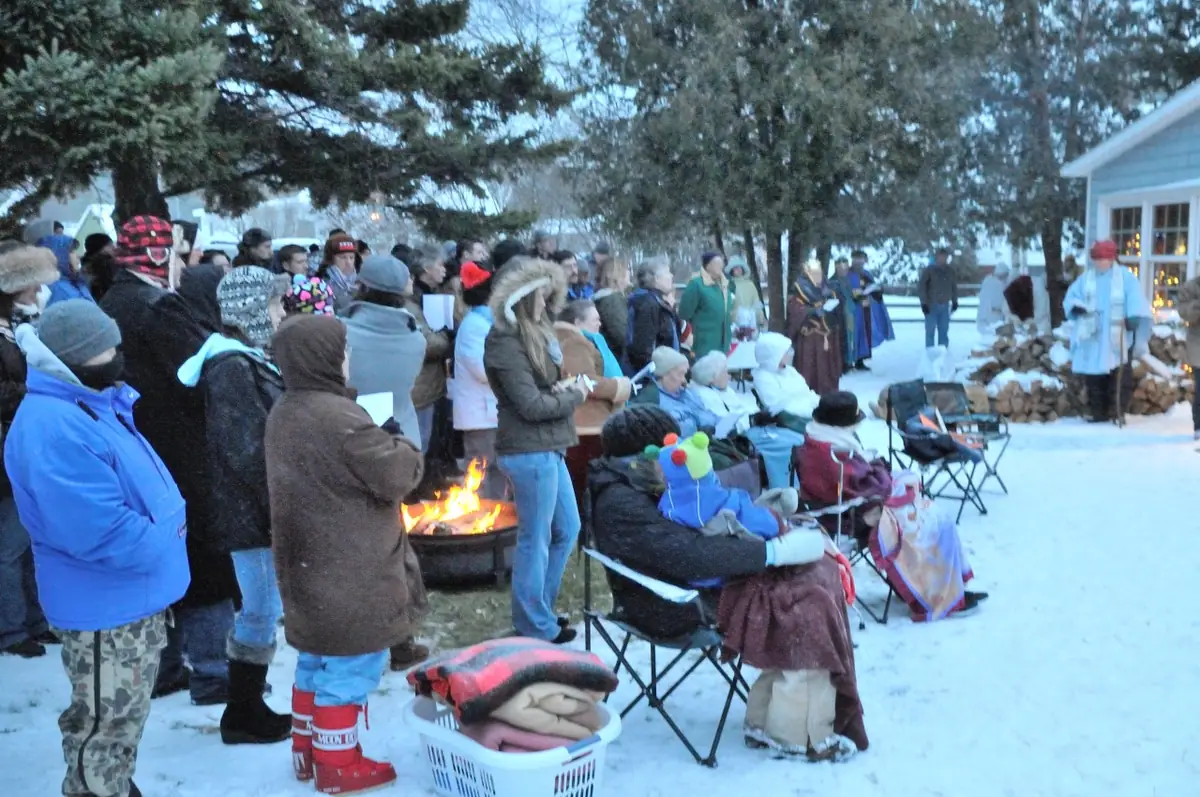 A bundled crowd gathers around a fire and costumed participants in a snowy outdoor nativity scene.