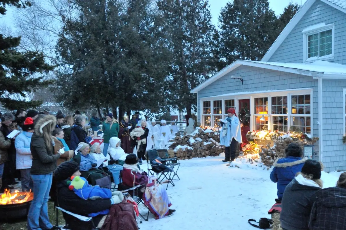 A winter outdoor gathering beside a shingled house, with costumed participants, seated spectators, and a fire.