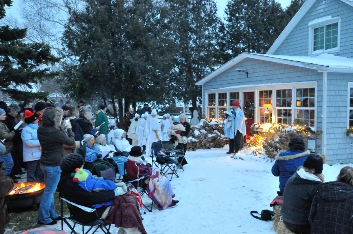 People gather in winter snow outside a house for a live nativity, with white-robed participants and a fire.