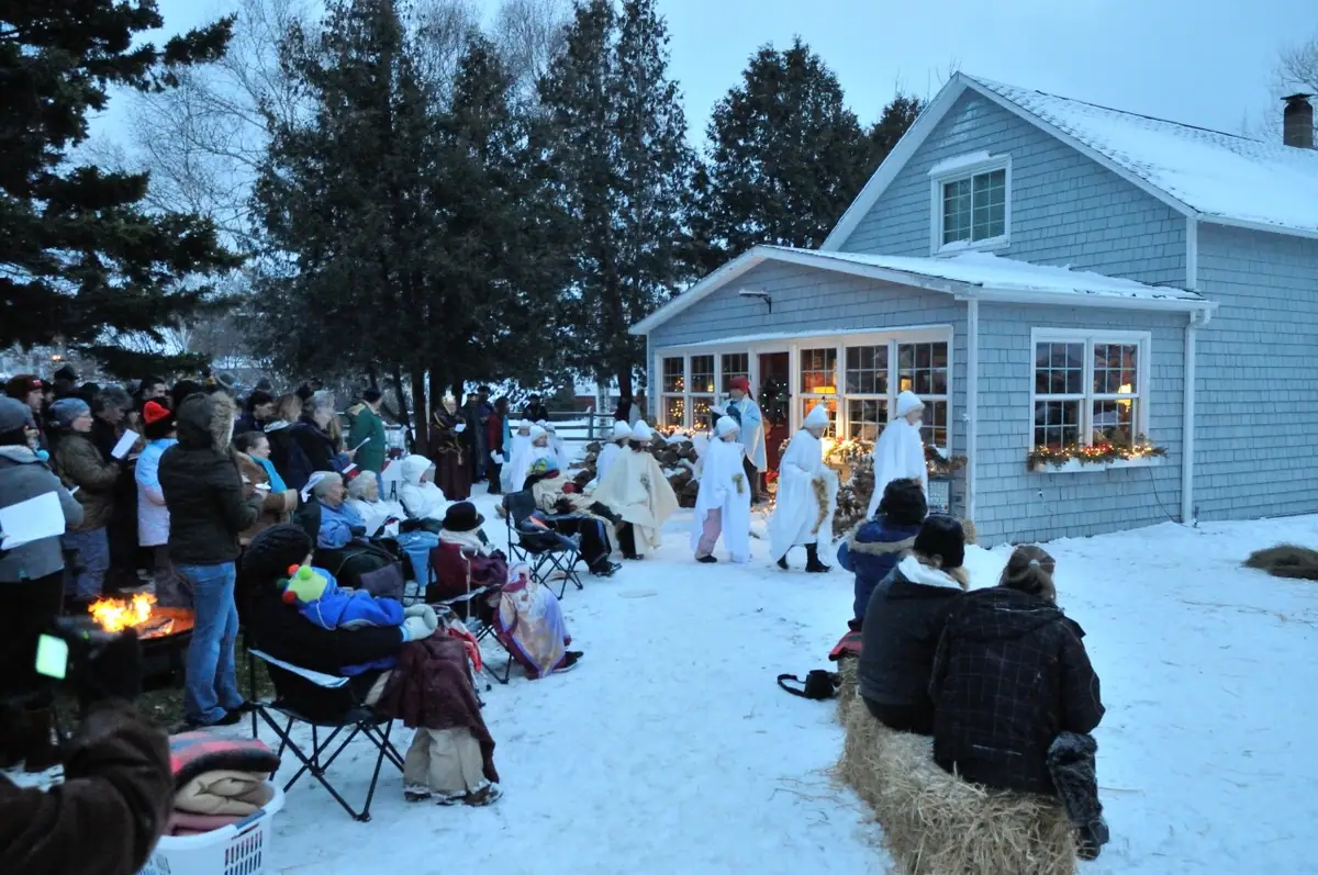 People gather in the snow as a robed procession passes a decorated house beside a small fire.