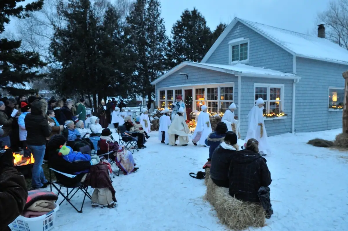 A winter nativity procession passes seated spectators beside a lit shingled house.