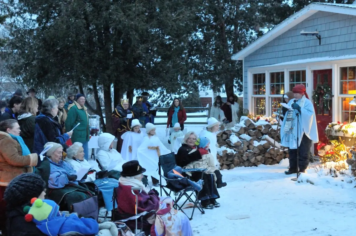Adults and children gather in snow outside a decorated house, with several children in white robes.