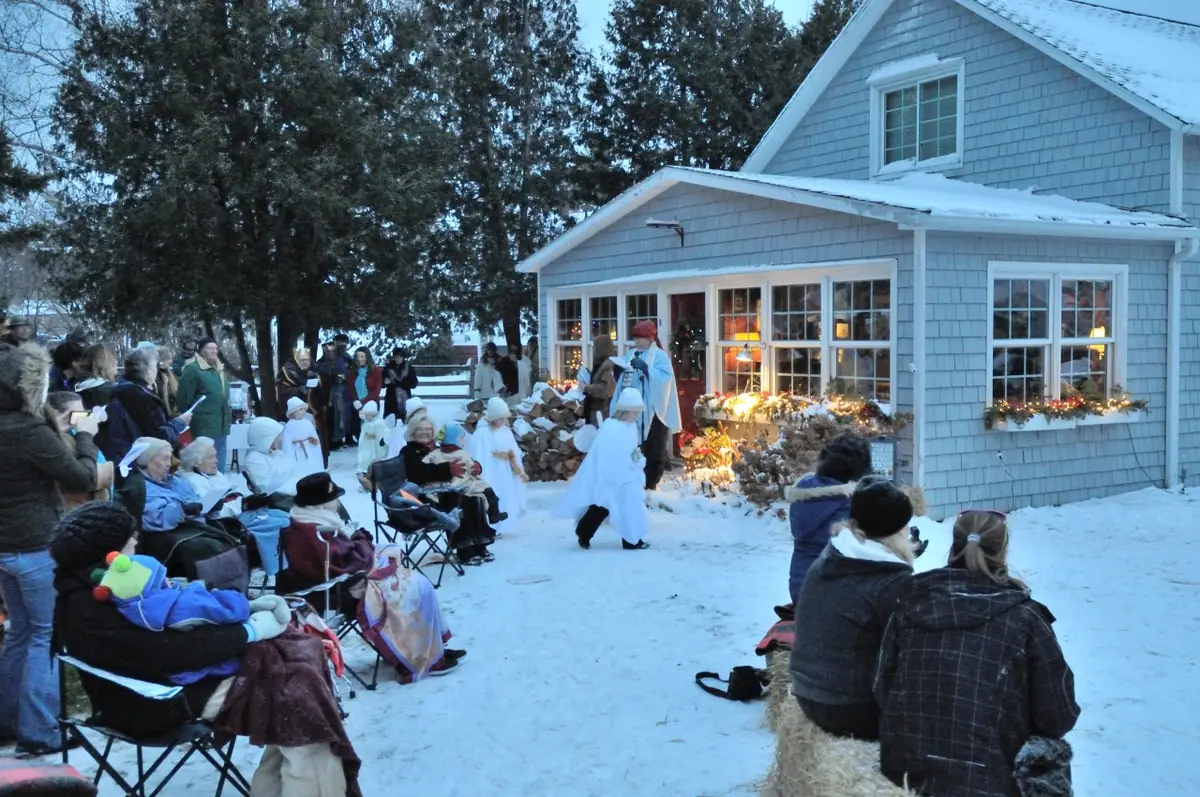 People watch a costumed outdoor performance in snowy surroundings beside a decorated shingled house.