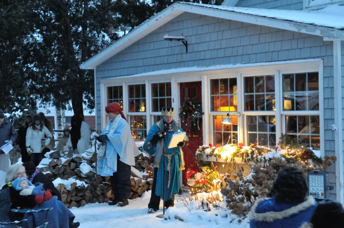 Costumed performers stand in snow before a shingled house as people watch.